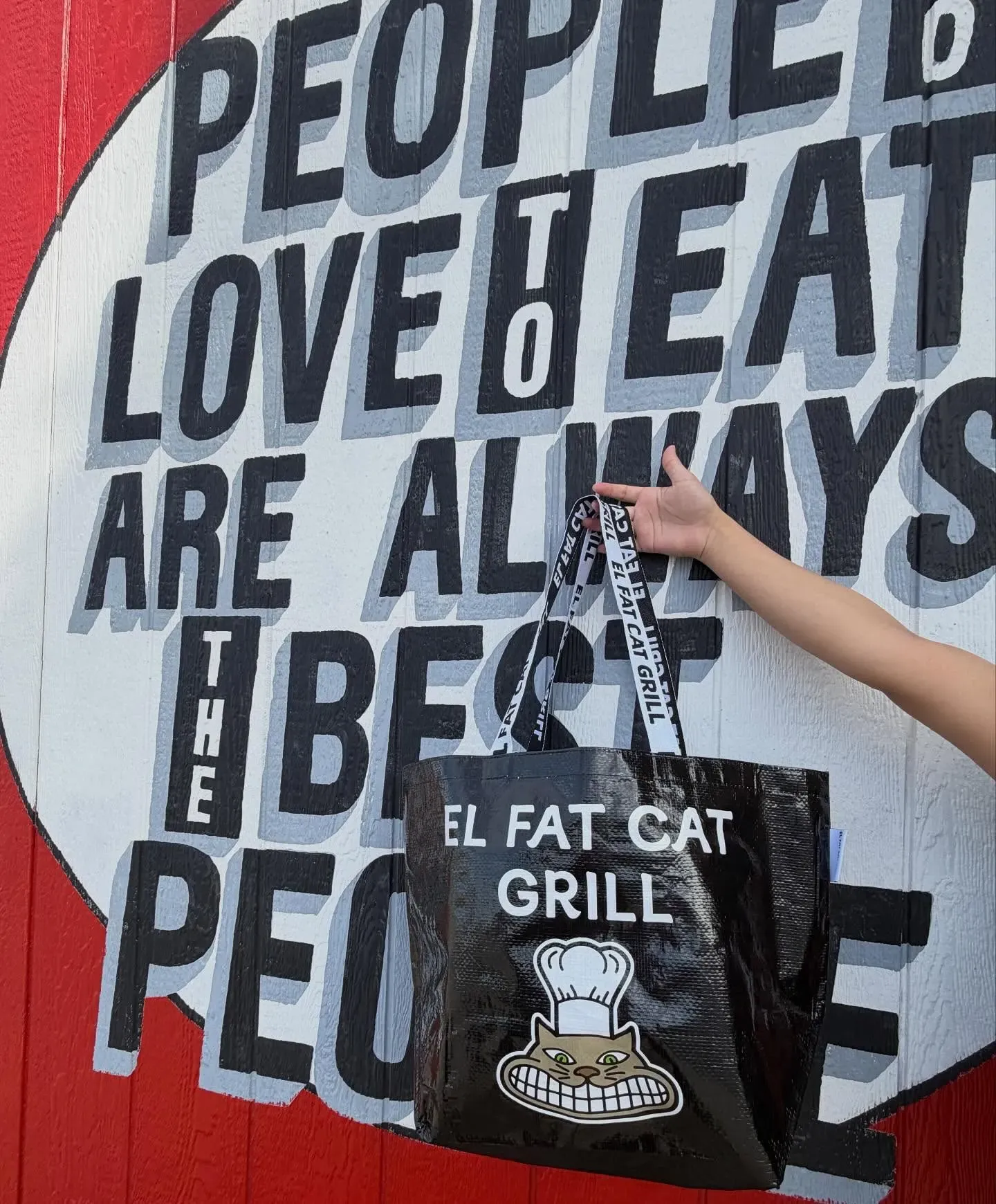 El Fat Cat Grill tote bag held up in front of the People Who Love To Eat Are Always The Best People mural